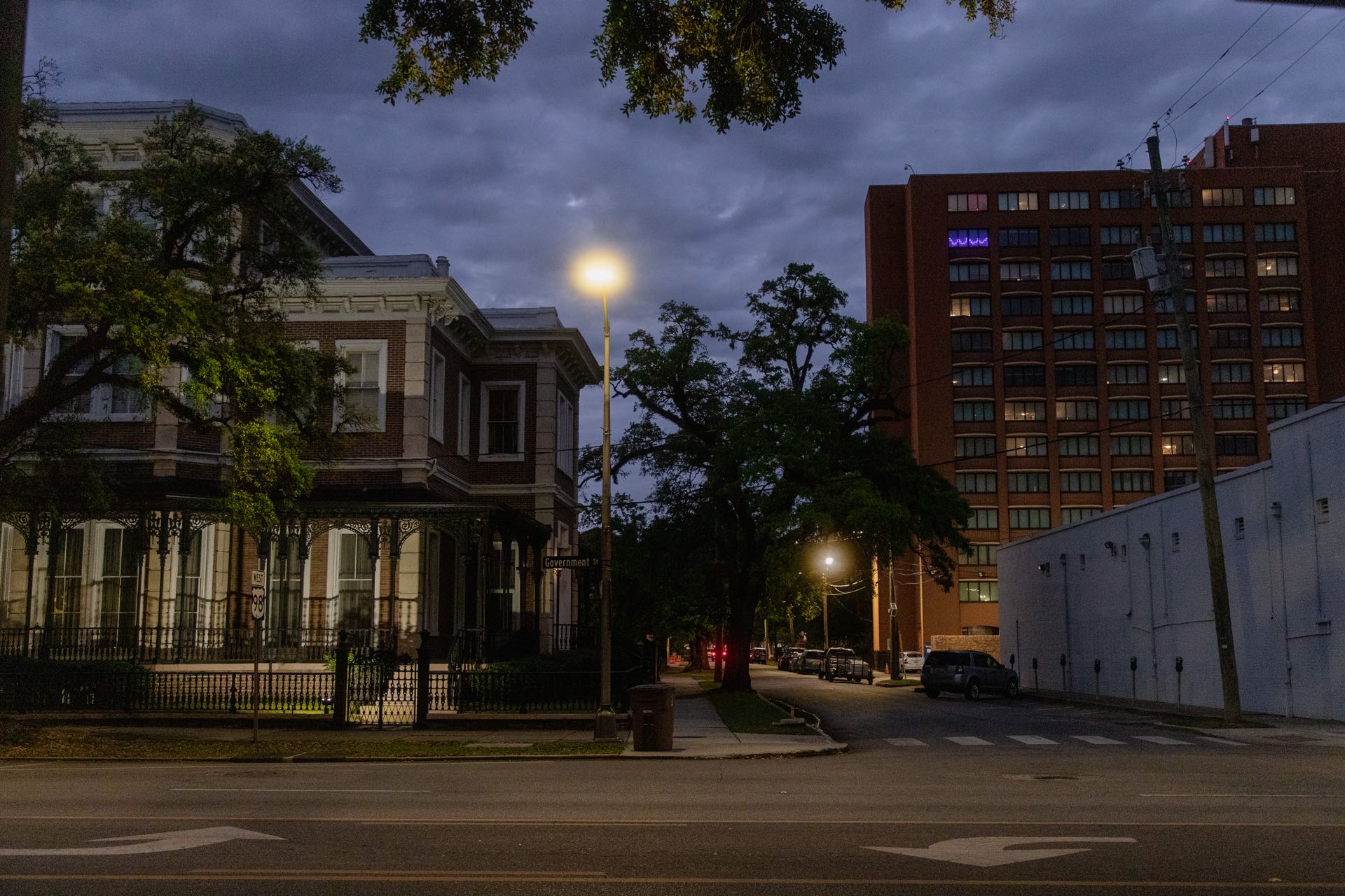 Government Street in downtown Mobile, Alabama at dusk — a historic iron-galleried building and a brick tower flanking an empty, lamp-lit street.