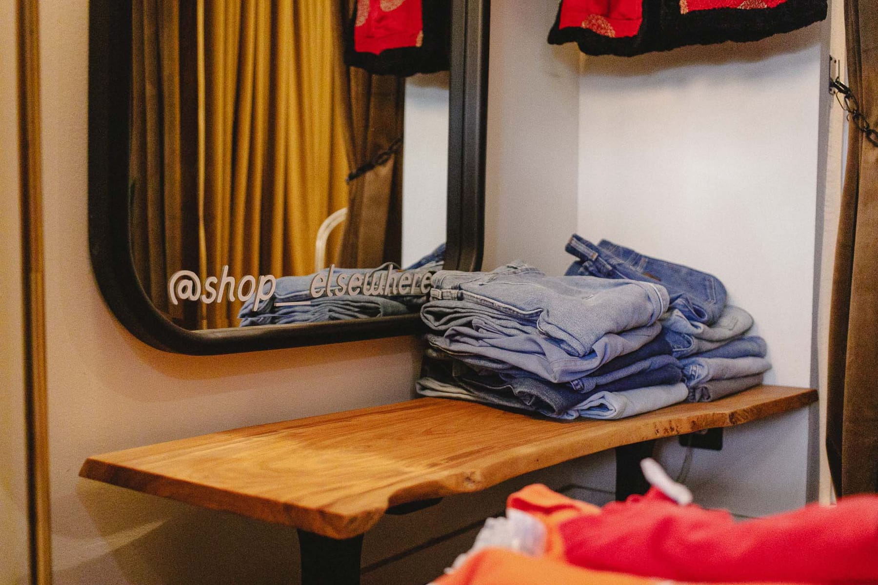 A live-edge wood bench by the fitting-room mirror, stacked with folded jeans. The mirror is lettered @shop__elsewhere.