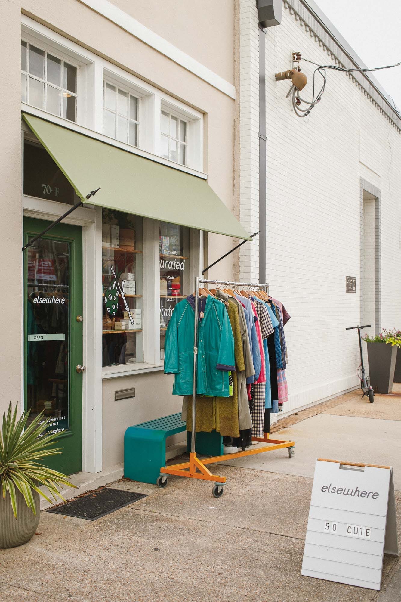 The shop from further down the sidewalk: green door under a pale green awning, a rolling rack of coats outside it, and an A-frame sign reading elsewhere, so cute.