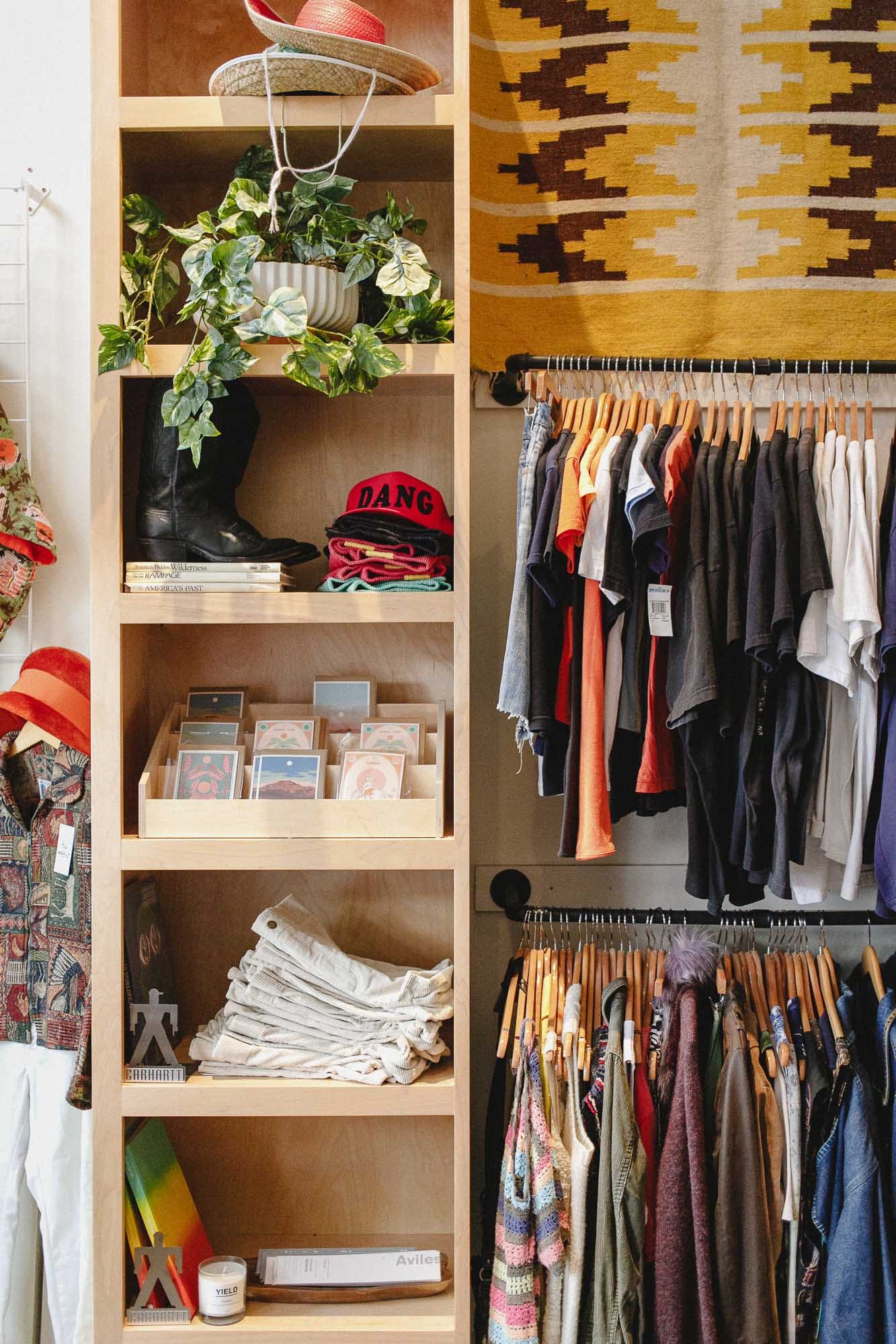 A tall built-in shelving bay: a straw hat, a trailing pothos, cowboy boots, a stack of books, a tray of art cards, folded textiles and a candle — racks of clothing to the right of it.