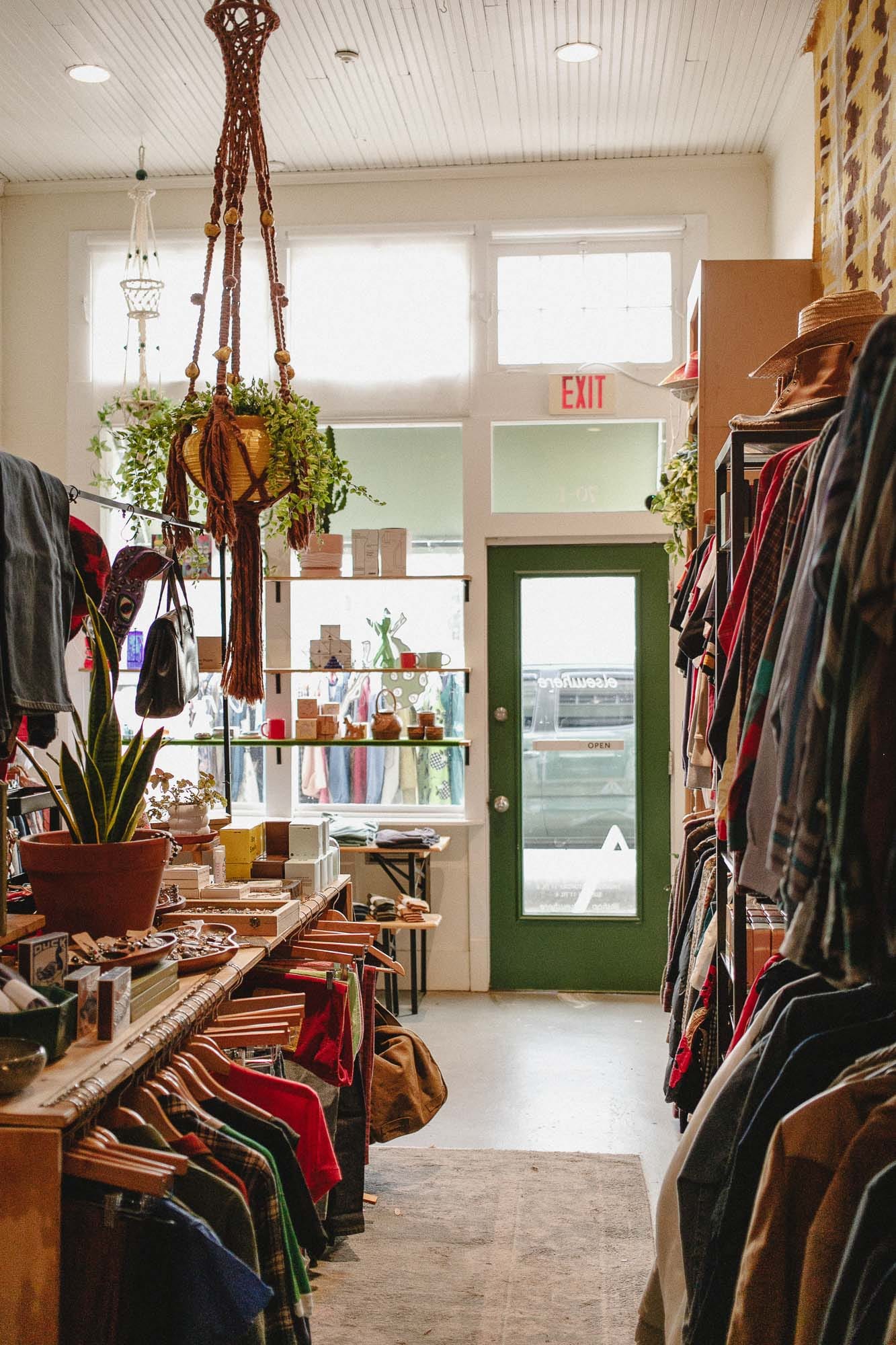The interior looking toward the green back door, clothing racks down the right wall, a long counter of goods on the left, a macramé planter hanging overhead.