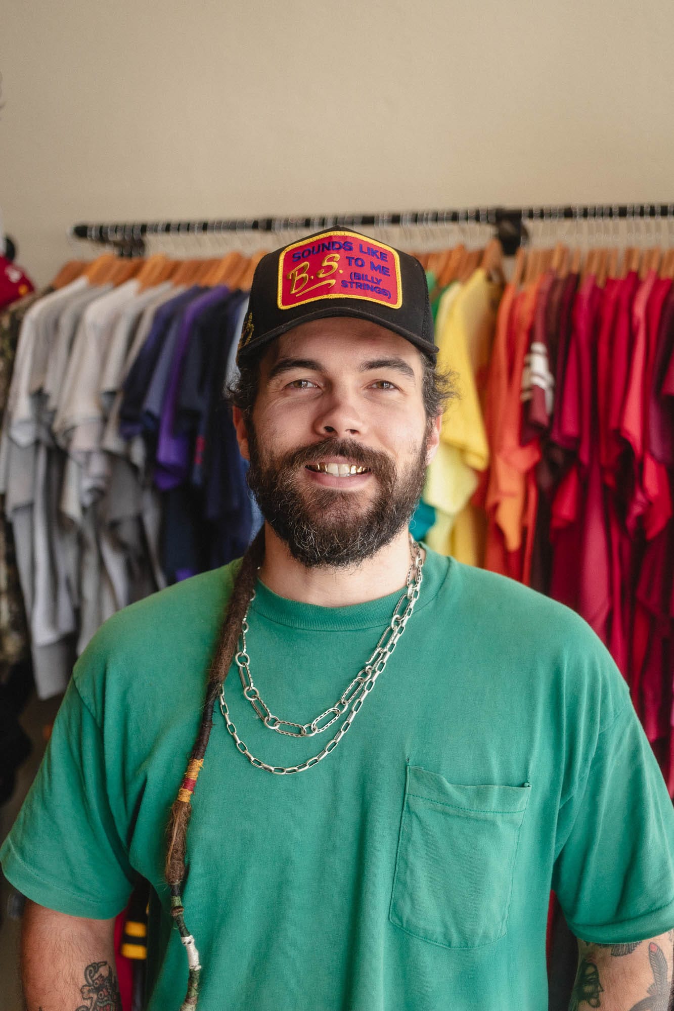 Sean in a green pocket tee and a black cap, smiling, in front of a rack of vintage T-shirts hung in color order.