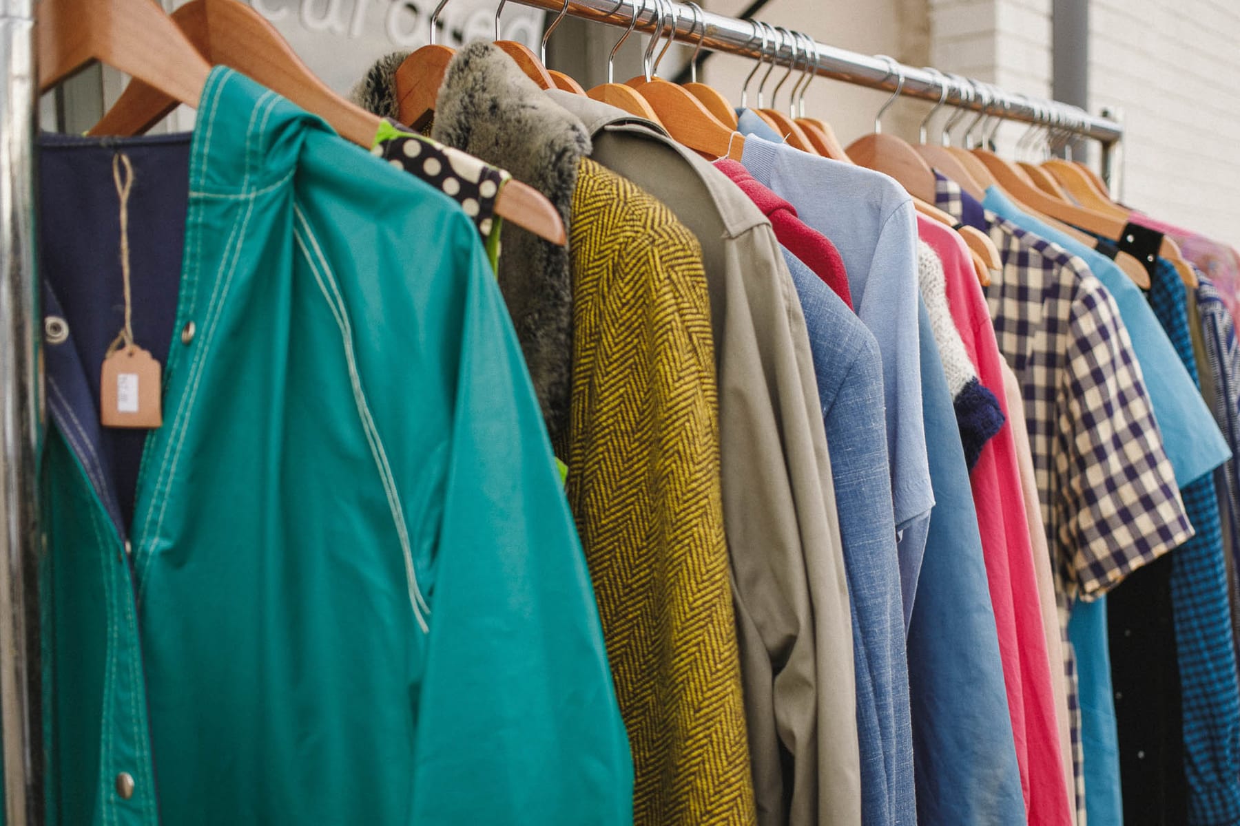 A rack of vintage clothing shot close along its length — a teal jacket, a mustard herringbone coat, checked and striped shirts on wooden hangers.