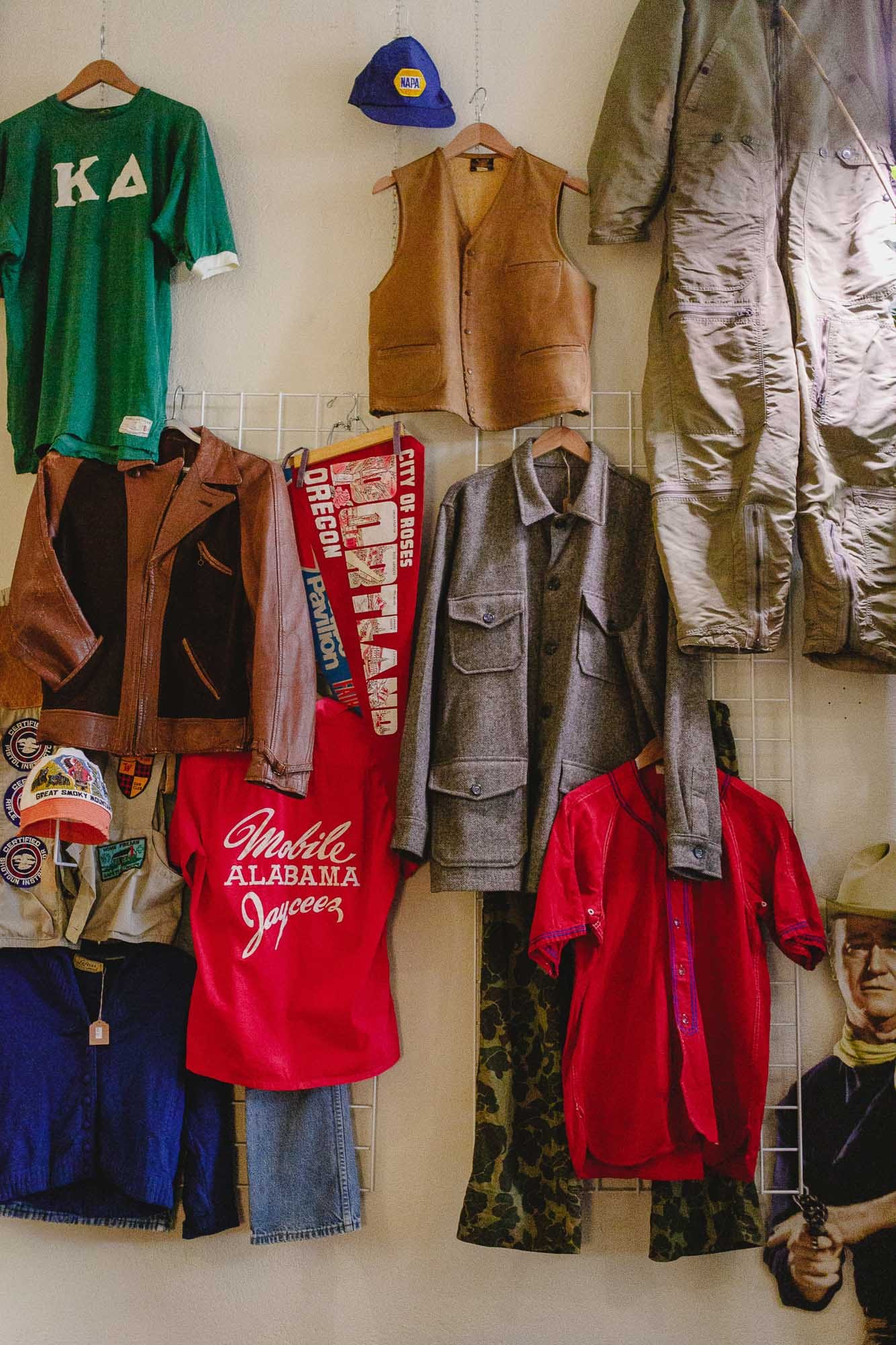 A wall of pieces hung on wire grid — a leather vest, a grey wool shirt jacket, camouflage trousers, a Portland pennant, and a red shirt lettered Mobile Alabama Jaycees.
